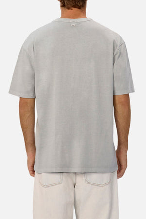 A person is seen from the back wearing an oversized Industrie The Del Sur Tee in light gray heavyweight fabric, paired with off-white pants featuring visible back pockets, standing against a plain white background.