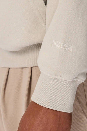 A close-up of a person's arm wearing the Industrie The Del Sur Washed Hoodie in light beige with "INDUSTRIE" embroidered on the sleeve, paired with tan pleated pants. The oversized fit offers comfort, and their hand rests relaxed at their side.