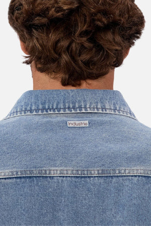 A person with curly brown hair is shown from behind wearing the Industrie The Denim Overshirt in light blue, featuring a small "Industrie" label on the collar, with a relaxed fit and vintage-inspired style.