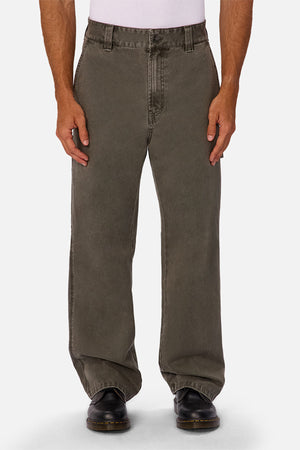 A person wears loose-fitting, faded olive green Industrie The Detroit Carpenter Pant in 100% cotton canvas, a tucked white shirt, and black shoes, standing against a white background for a classic street style look.