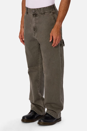 Someone wearing loose-fitting, faded olive-green Industrie The Detroit Carpenter Pant in 100% cotton canvas, a white shirt, and black leather boots stands with arms relaxed at sides against a plain white background for a street style look.