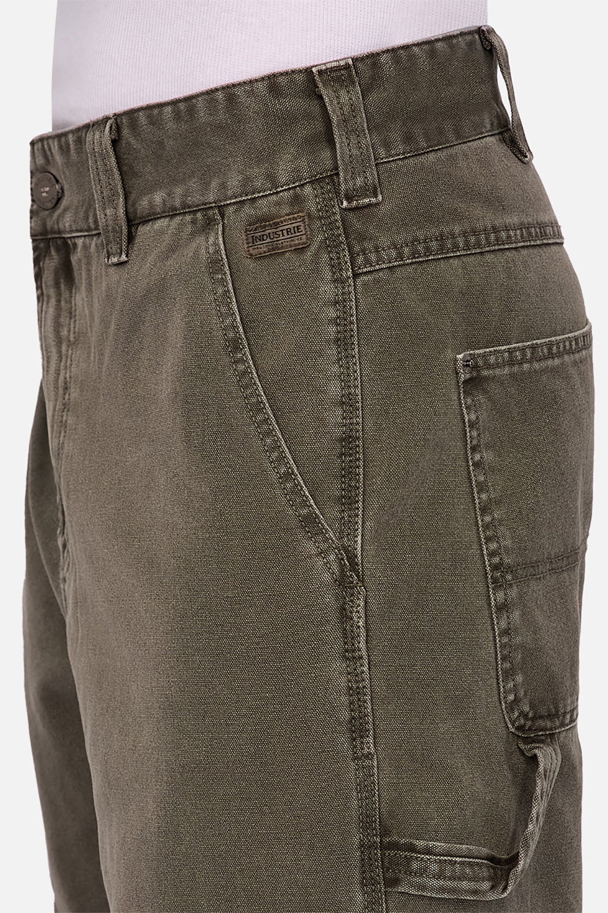 Close-up of a person wearing Industrie The Detroit Carpenter Pant in olive green 100% cotton canvas, with visible stitching, belt loops, button closure, and multiple pockets. Paired with a tucked-in white shirt for a sharp street style look.