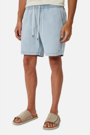A person is dressed in a casual outfit comprising Industrie The Dublin Short in light blue, featuring an elasticised waistband, paired with a white top and beige slide sandals. This ensemble provides a breathable fit against the plain white background.