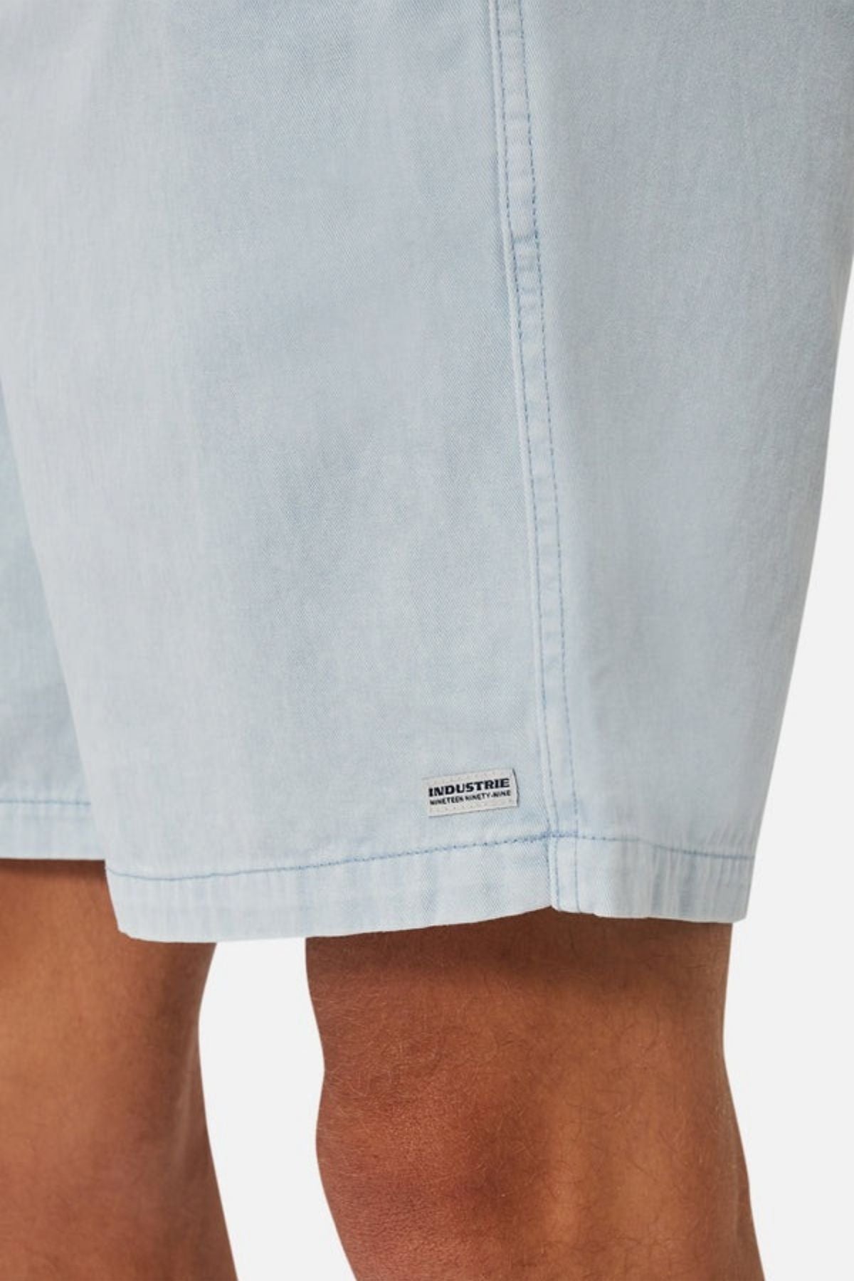 Close-up of a person's legs wearing the Industrie The Dublin Short in light blue denim. A small label near the hem reads "INDUSTRIE." The image showcases the fabric's texture, color, and breathable fit.