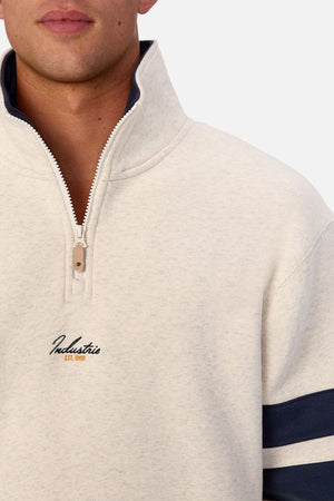 Wearing the Industrie The East Village Track Top in light beige, featuring dark blue sleeve stripes and "Industrie Est. 1999" embroidered on the chest. Its relaxed fit enhances casual style; the face is partially visible.