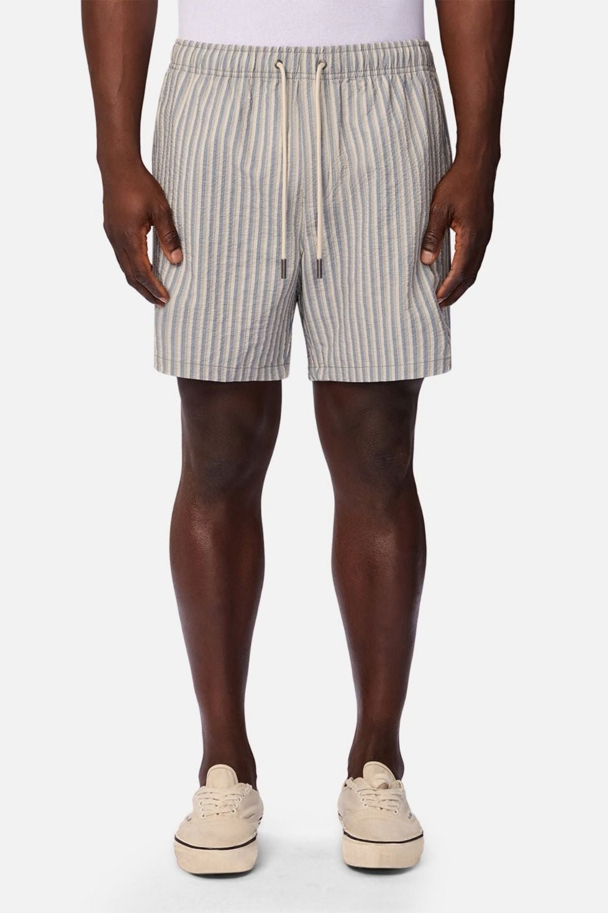 A person wearing a white tank top, beige sneakers, and the Industrie The Formosa Swim Short—featuring light blue and beige stripes with a drawstring—stands facing forward against a plain background.