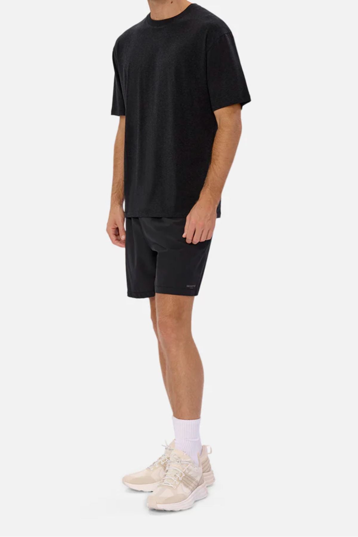 A man wears an Industrie The Freeman Athletic Tee in black, black shorts, white crew socks, and beige sneakers while standing against a plain white background. His head is not visible in the image.
