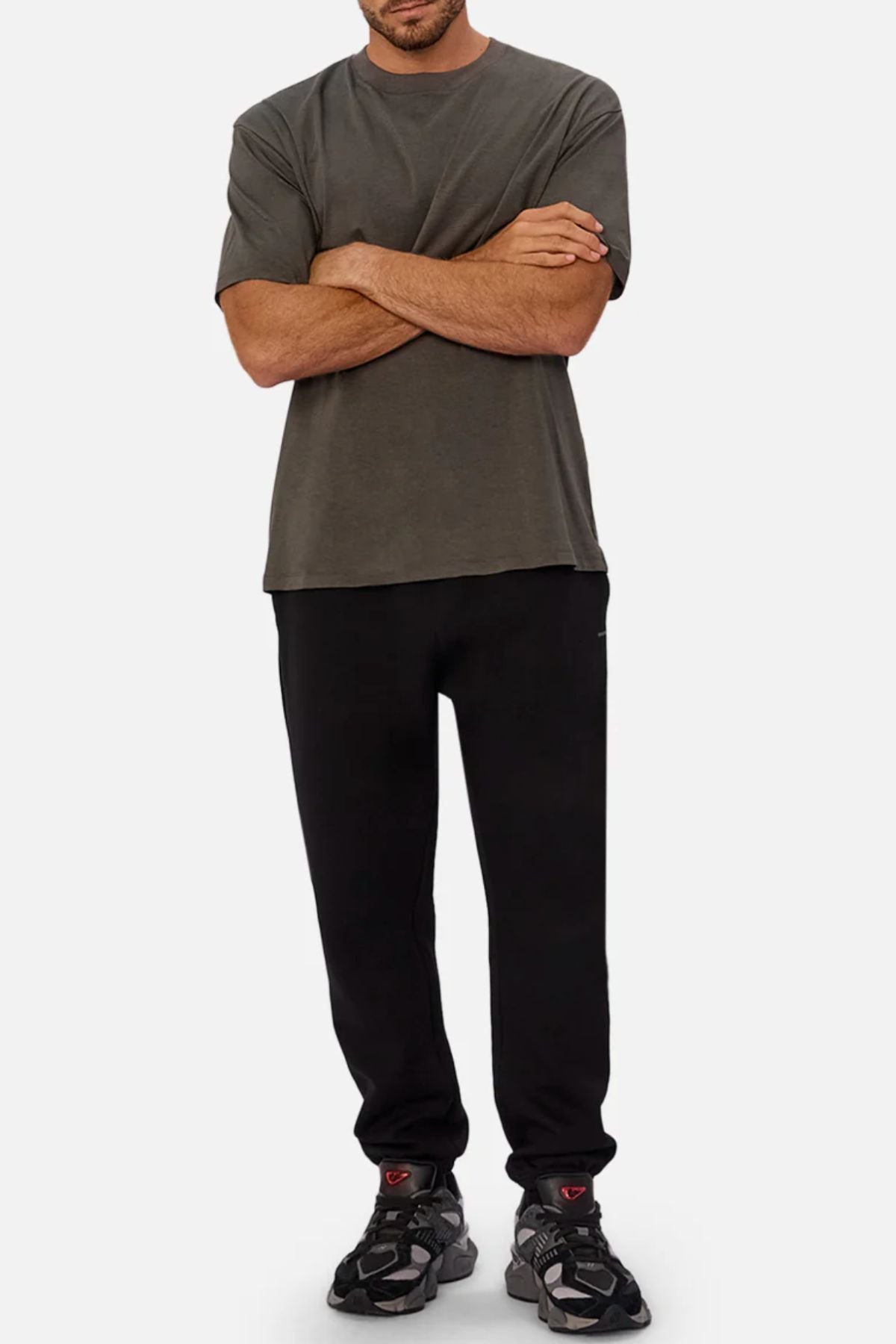 A man stands with arms crossed, wearing the Industrie The Freeman Athletic Tee in dark gray, paired with black sweatpants and black athletic sneakers. The background is plain white, and his face is partially out of frame.