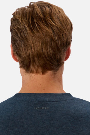 A person with short light brown hair is seen from the back wearing the Industrie The Freeman Athletic Tee, a dark blue relaxed-fit top featuring "INDUSTRIE" printed on the upper back. The background is plain and light-colored.