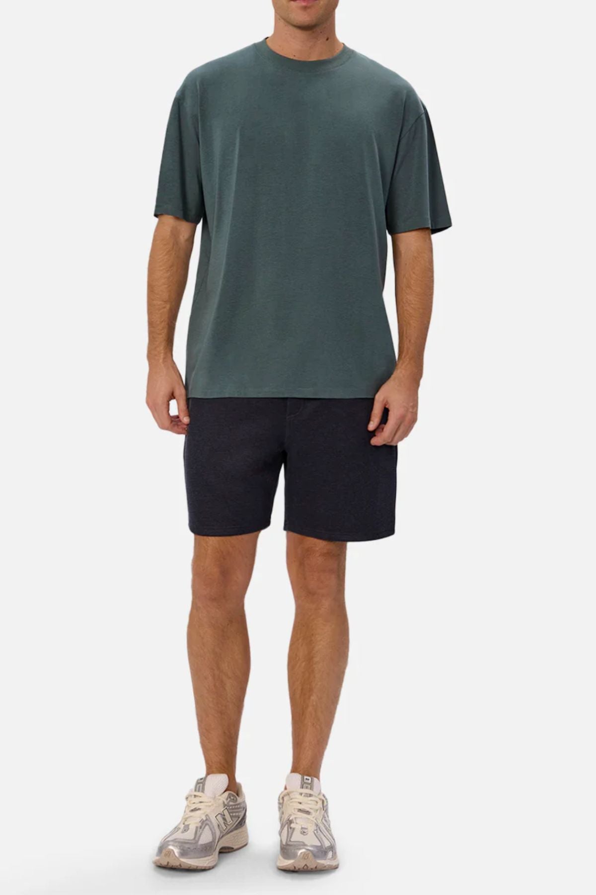 A man wears the Industrie The Freeman Athletic Tee in a relaxed fit, dark green color, paired with dark shorts and light athletic sneakers. He stands against a plain white background with his head not visible in the image.