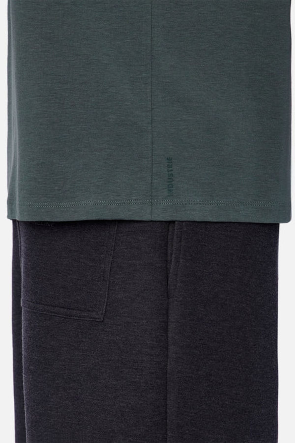 A close-up of a fabric detail showcases the bottom of the dark green Industrie The Freeman Athletic Tee, layered over dark gray sweatpants. The tee includes subtle branding on the hem, and the sweatpants feature a visible side pocket.
