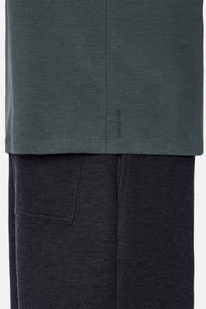 A close-up of a fabric detail showcases the bottom of the dark green Industrie The Freeman Athletic Tee, layered over dark gray sweatpants. The tee includes subtle branding on the hem, and the sweatpants feature a visible side pocket.