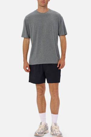 A man stands against a plain background wearing the Industrie The Freeman Athletic Tee in grey, paired with black shorts, white socks, and white sneakers.