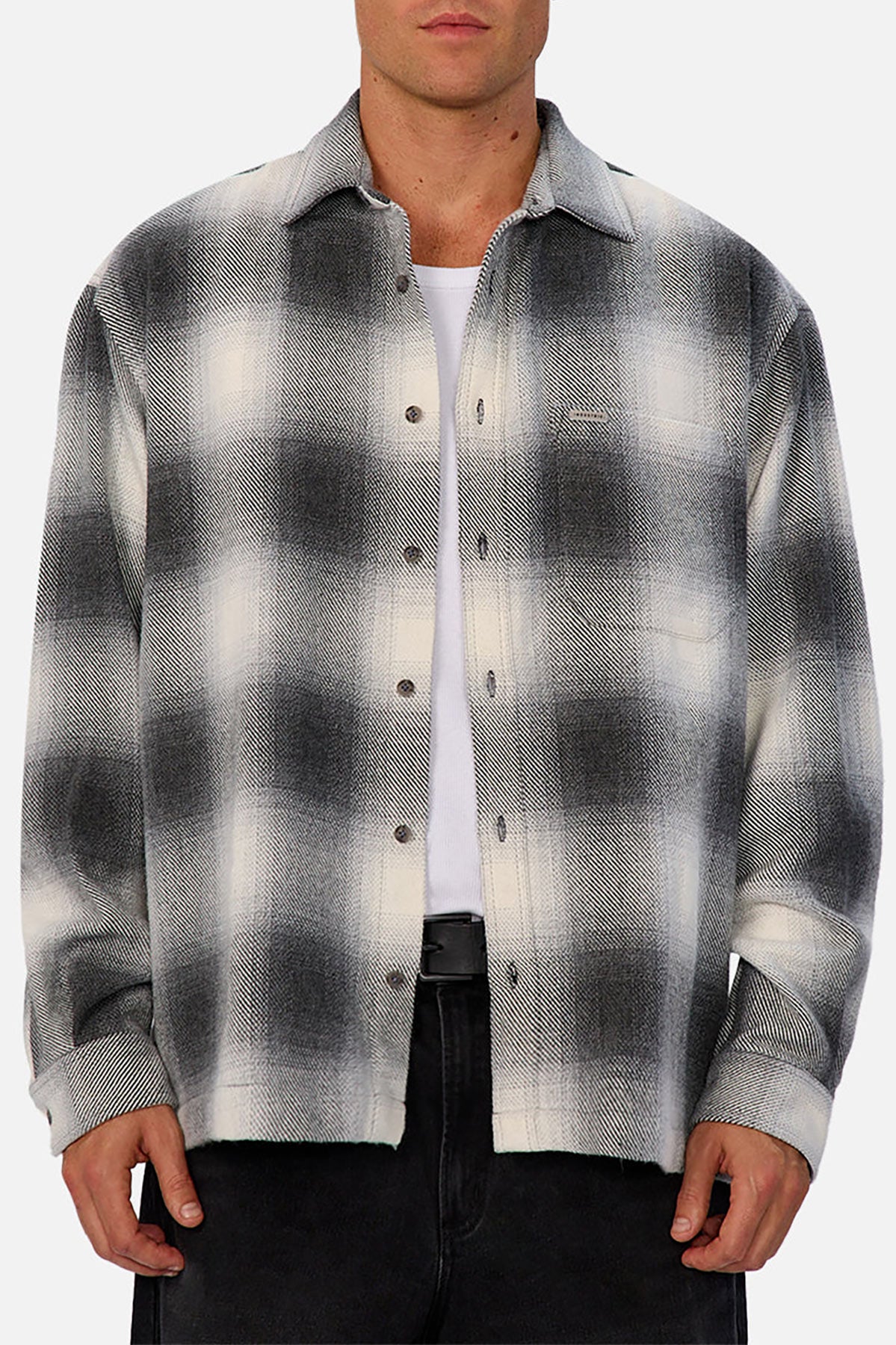 A person wears the Industrie The Highland Shirt in black and white shadow check, untucked with the top button undone, over a white T-shirt and black pants, standing against a light gray background, highlighting its tailored fit.