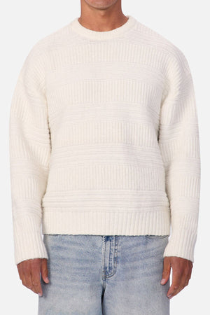 Someone wearing the Industrie The Huntington Knit in off-white with a textured pattern and light blue jeans stands against a plain light background.