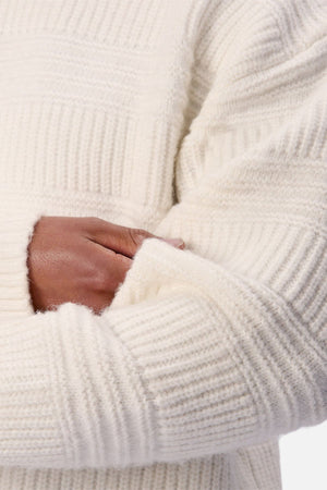 Someone wearing the Industrie The Huntington Knit, a chunky white crew neck sweater, crosses one arm over their body with their hand tucked into the opposite sleeve.