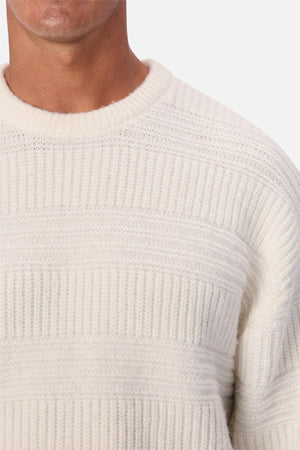 A close-up of a person wearing the Industrie The Huntington Knit, featuring a textured, off-white ribbed pattern. Only the lower half of the face, neck, and upper torso are visible.