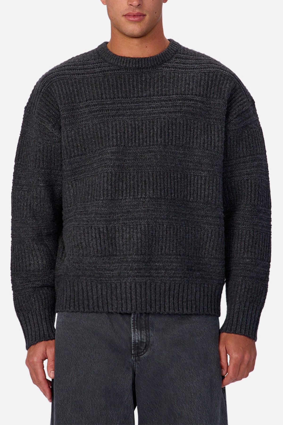 A person wearing the Industrie The Huntington Knit, a textured dark gray crew neck sweater with long sleeves, paired with faded black jeans and standing against a plain light background.