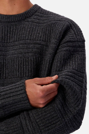A person is shown from the shoulders down wearing the Industrie The Huntington Knit, a regular fit dark gray textured crew neck sweater, gripping their left arm above the elbow against a plain white background.