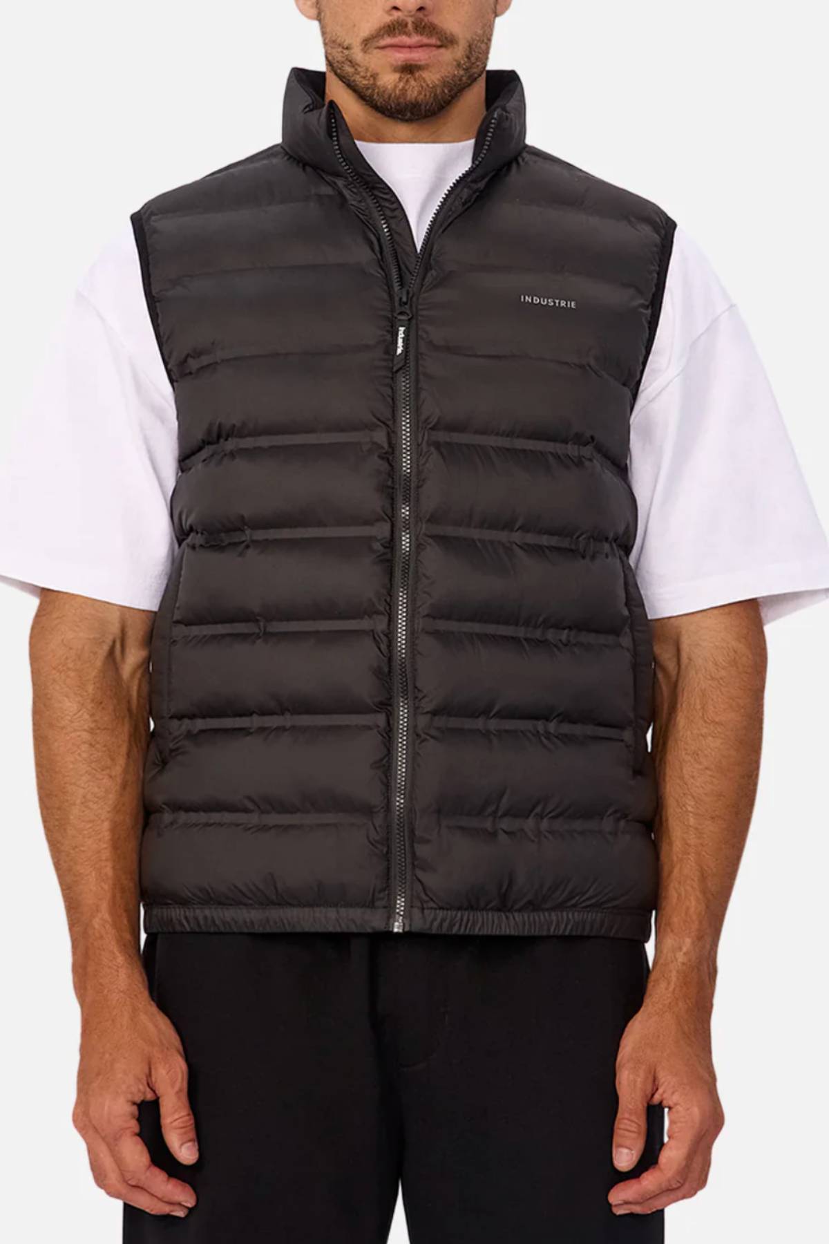 A man wears the Industrie The Kyoto Puffer Vest in black, featuring a heat-sealed design, front zipper, and subtle left-chest branding, over a white short-sleeve tee and black pants, standing against a plain light background.