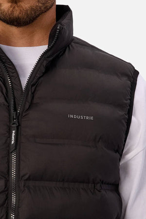Close-up of a person wearing the Industrie The Kyoto Puffer Vest, featuring a black heat-sealed design with "INDUSTRIE" on the chest, layered over a white shirt. Only the lower face and upper torso are visible.