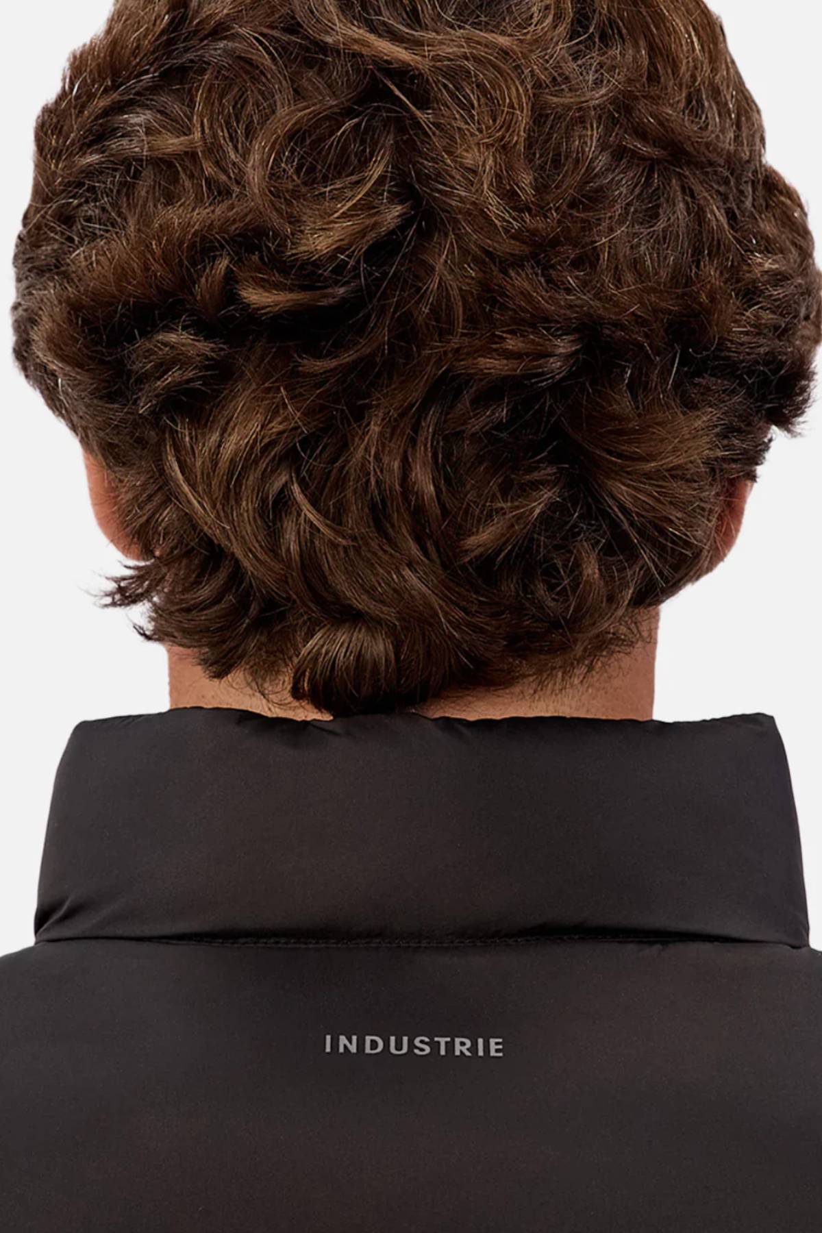 A close-up of the back of a person with short, wavy brown hair, wearing the Industrie The Kyoto Puffer Vest with "INDUSTRIE" on the collar and internal pockets for added functionality.