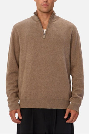 A person wears the Industrie The Lakewood Zip Neck Knit—a light brown cotton jumper with a small collar, relaxed fit, and rib knit cuffs—over a white undershirt and loose dark pants, standing against a plain light background.