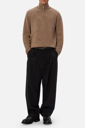 A person wearing the Industrie The Lakewood Zip Neck Knit in light brown, loose black pleated trousers, a black belt, and black shoes stands against a plain white background with hands in pockets.