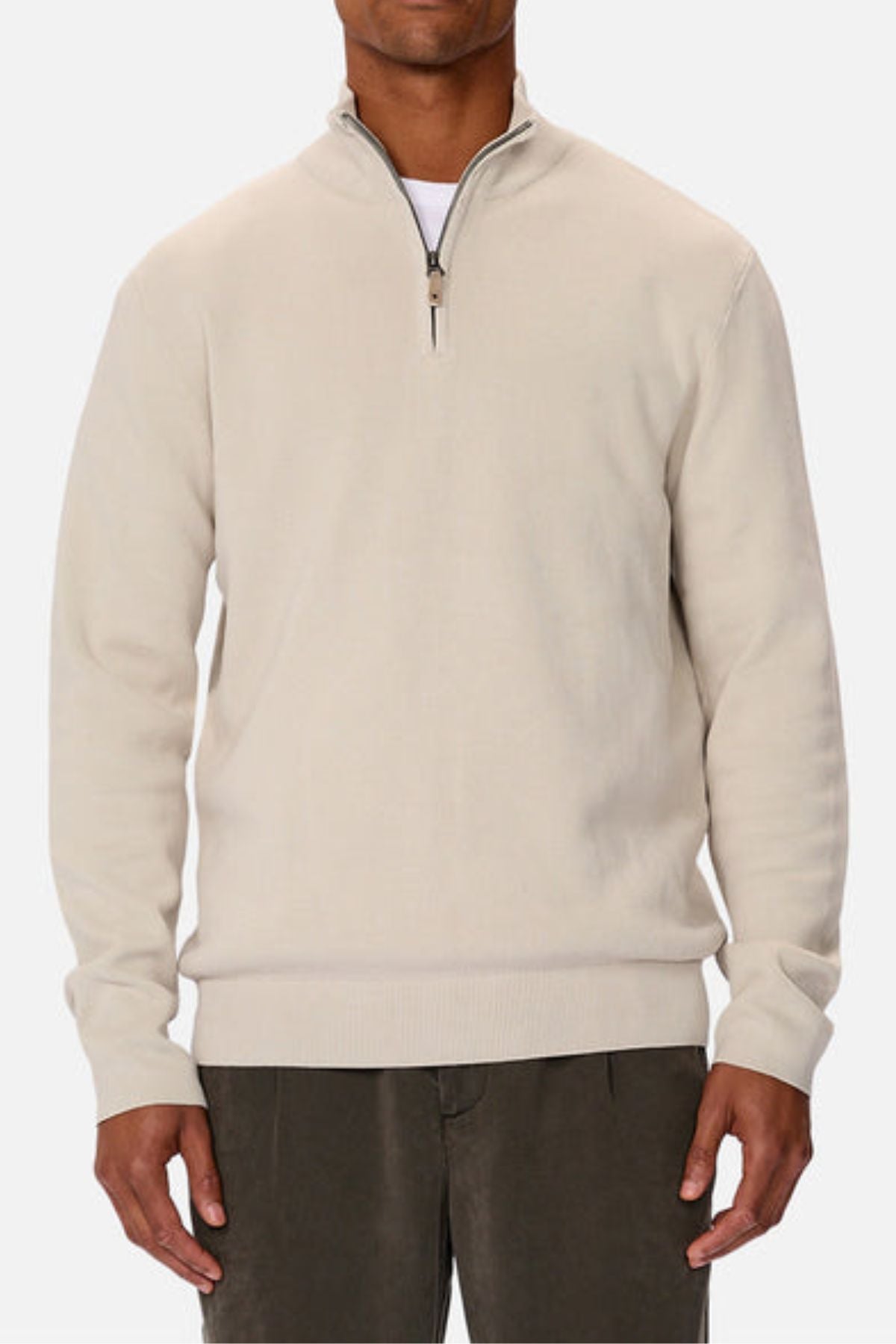 A man wears the Industrie The Lakewood Zip Neck, a light beige cotton jumper with long sleeves and rib knit cuffs, paired with dark gray pants, standing against a plain white background.