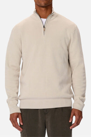 A man wears the Industrie The Lakewood Zip Neck, a light beige cotton jumper with long sleeves and rib knit cuffs, paired with dark gray pants, standing against a plain white background.