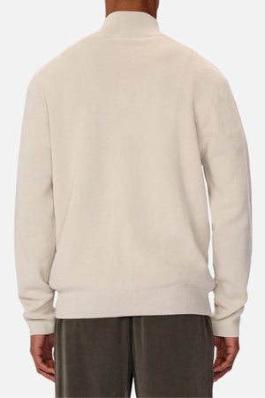 A person is shown from the back, standing against a plain white background, wearing the Industrie The Lakewood Zip Neck—a long-sleeved, light beige cotton jumper with rib knit cuffs—and dark pants.