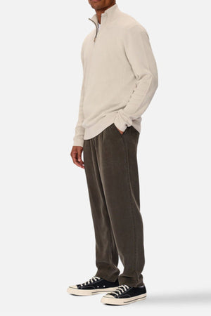 A man stands with his hands in his pockets, wearing the Industrie The Lakewood Zip Neck jumper in light beige, paired with loose dark gray pants and black low-top sneakers with white soles against a plain white background.
