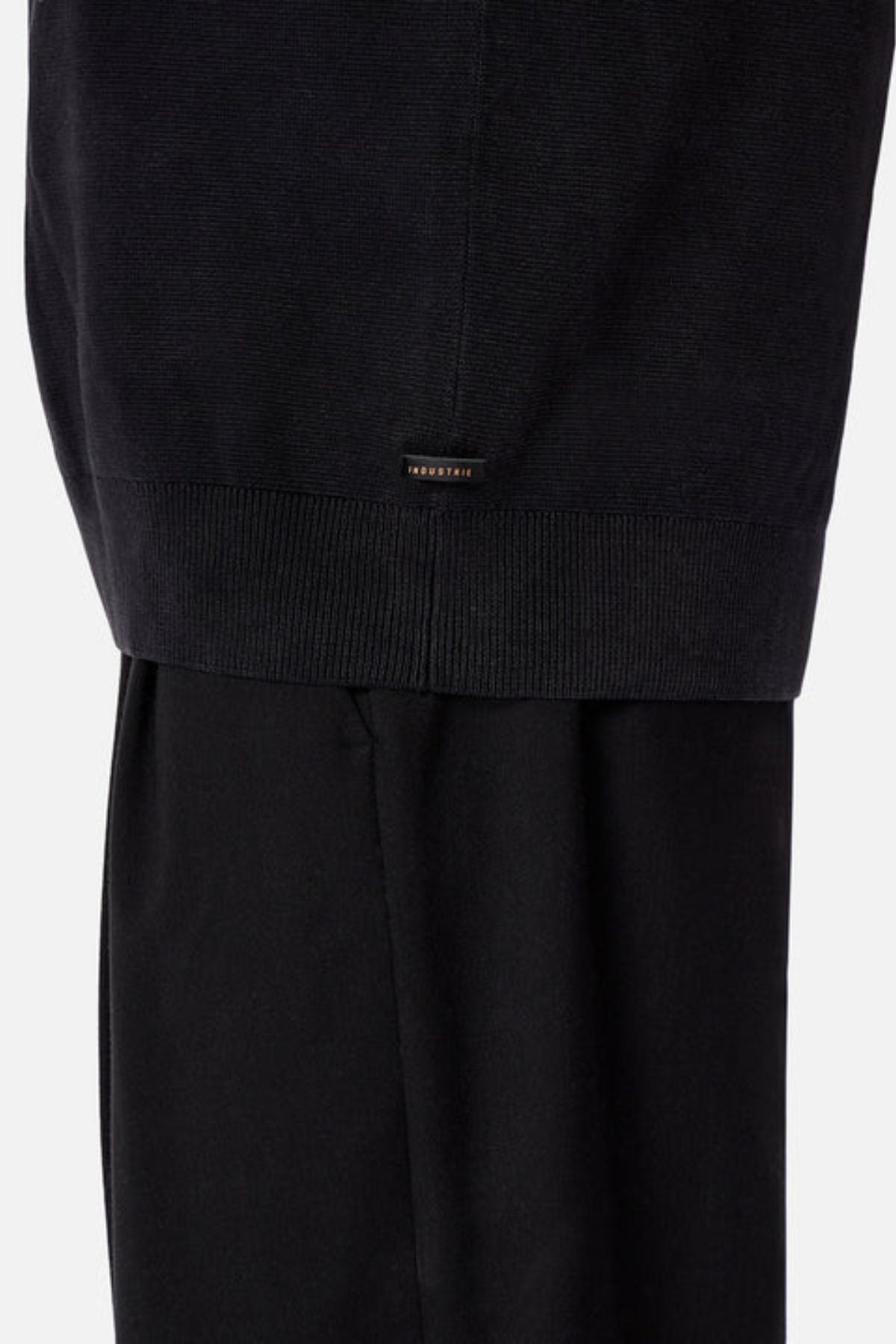 Close-up of the lower part of a person wearing Industrie The Lakewood Zip Neck and black pants, highlighting the ribbed hem and a small rectangular label above it against a plain, light-colored background.