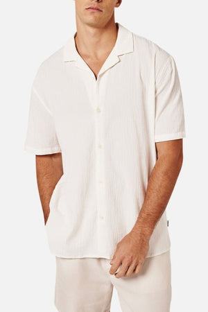 A man wearing the Industrie The Magna Short Sleeve Shirt—textured, white, slightly oversized, and breathable—paired with light-colored shorts stands with one hand in his pocket against a plain background.
