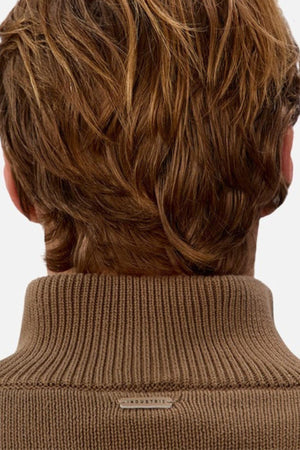 A close-up of the back of a person's head with light brown, wavy hair. They are wearing the Industrie The Maine Zip Neck Knit, a ribbed brown turtleneck with a small "INDUSTRIE" tag at the neck.