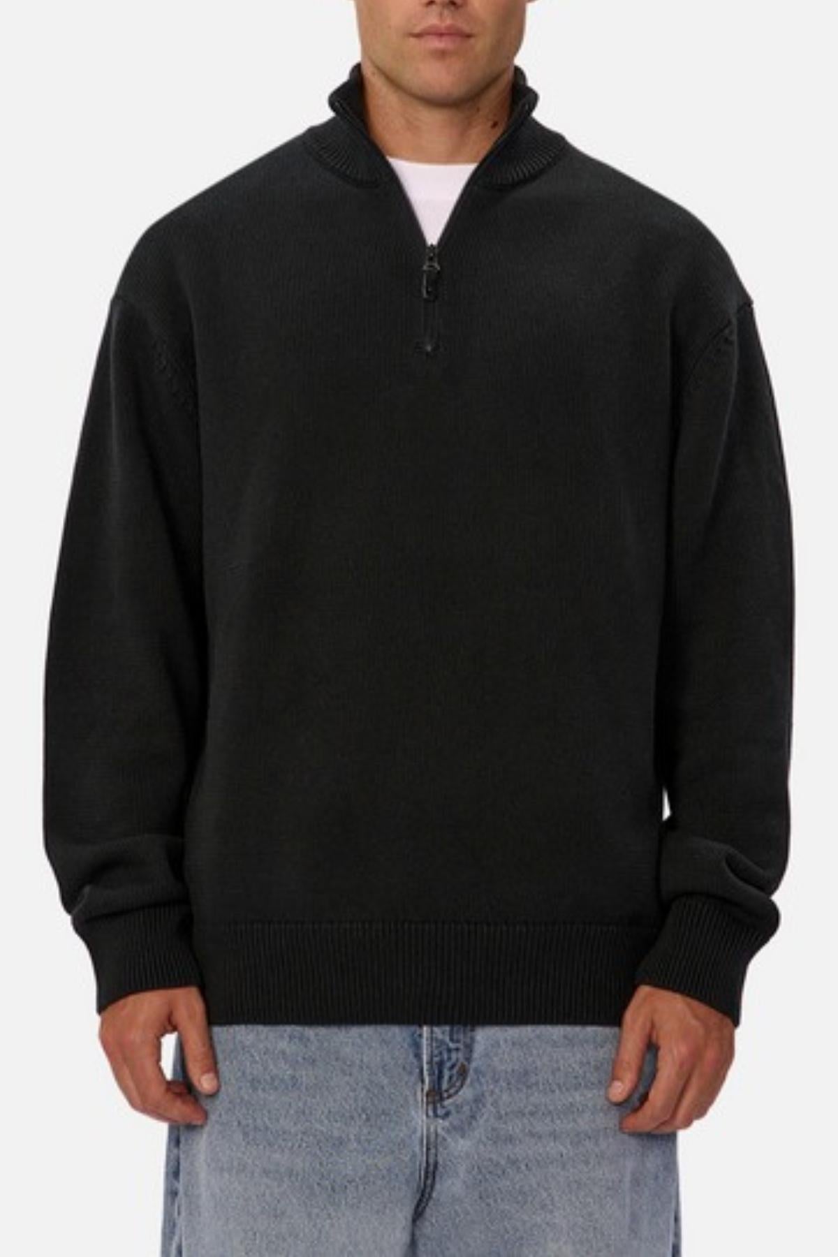 Wearing a black Industrie The Maine Zip Neck Knit, regular fit, heavyweight cotton, over a white shirt and light blue jeans, someone stands against a plain white background.
