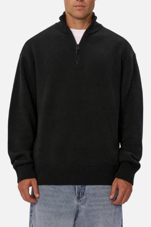 Wearing a black Industrie The Maine Zip Neck Knit, regular fit, heavyweight cotton, over a white shirt and light blue jeans, someone stands against a plain white background.