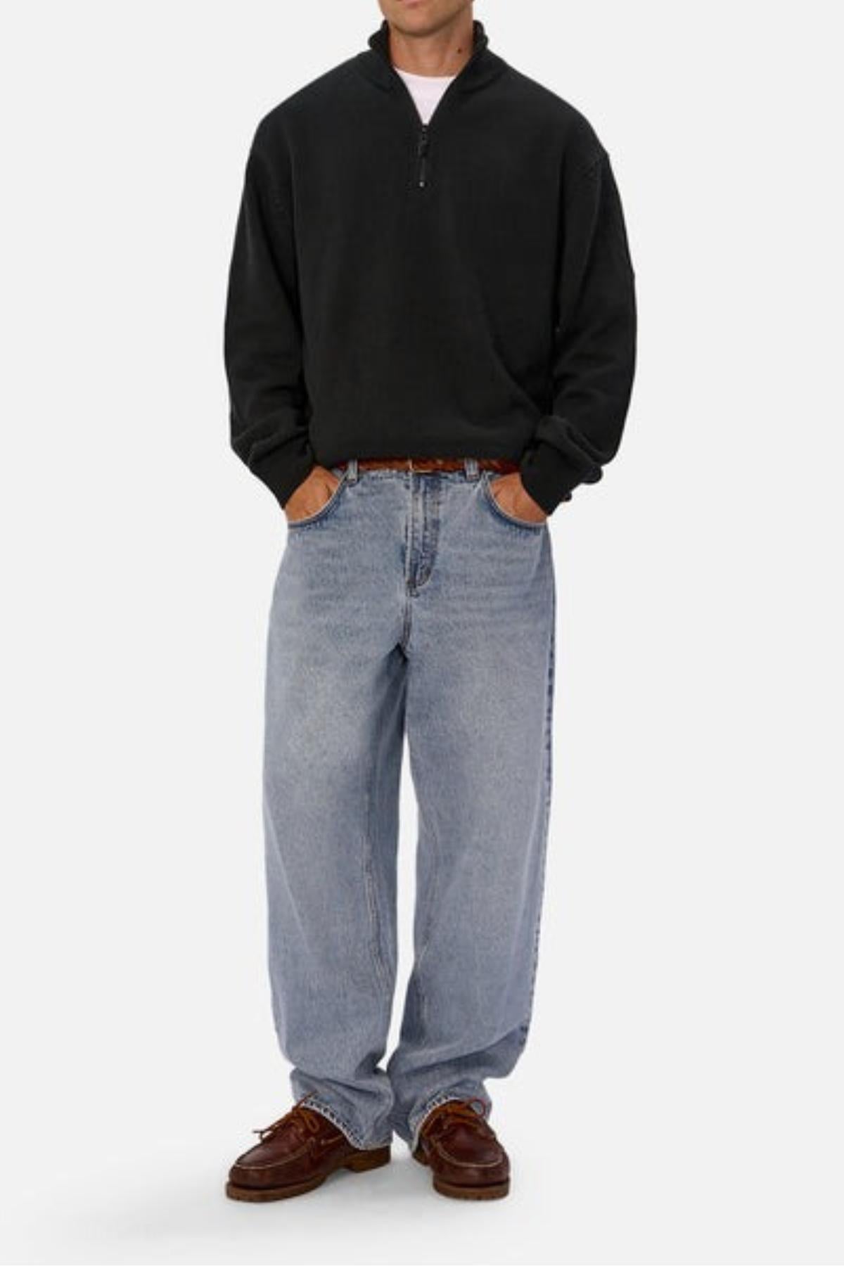 A person in a black Industrie The Maine Zip Neck Knit stands against a plain background, paired with light blue baggy jeans and brown boat shoes, hands in pockets.