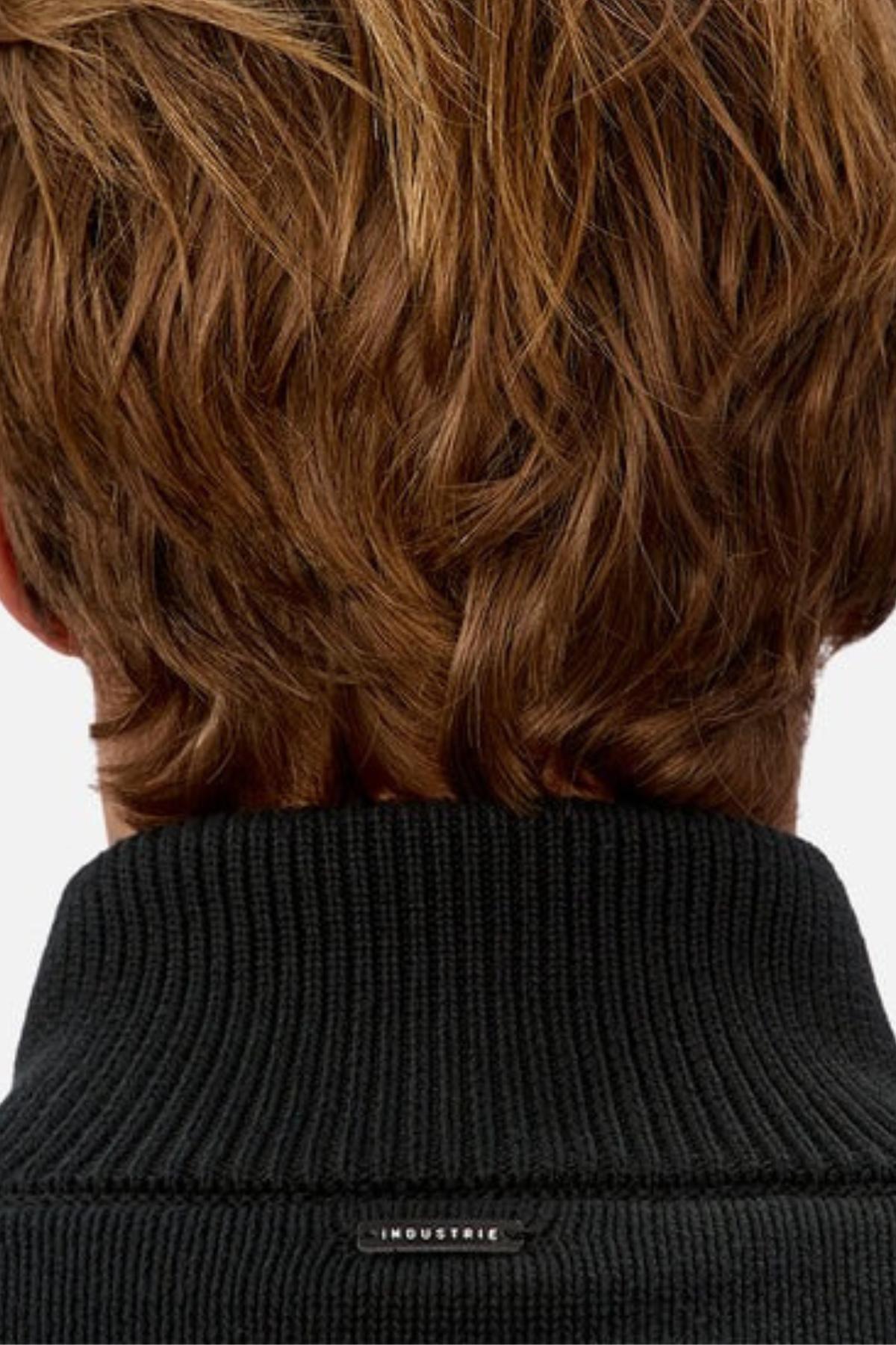 Close-up of the back of a person with short, wavy brown hair wearing the Industrie The Maine Zip Neck Knit, made from heavyweight cotton and detailed with a small "INDUSTRIE" label on its black ribbed collar.