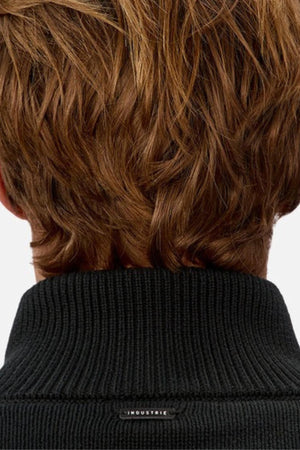 Close-up of the back of a person with short, wavy brown hair wearing the Industrie The Maine Zip Neck Knit, made from heavyweight cotton and detailed with a small "INDUSTRIE" label on its black ribbed collar.