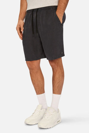 A man wears Industrie The Marina Linen Short in black with a drawcord waist, paired with a light gray shirt, white socks, and white sneakers. His left hand is in his pocket; only his body from the chest down is shown.