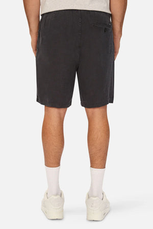 A person is shown from the back, standing against a plain white background, wearing a light gray t-shirt, Industrie The Marina Linen Short in a relaxed fit with drawcord waist, white crew socks, and white sneakers.