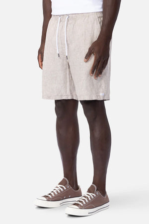 A person models the Industrie The Marina Linen Short in light beige, paired with a white shirt and brown low-top sneakers, standing against a plain background. The image highlights the breathable shorts from the waist down.