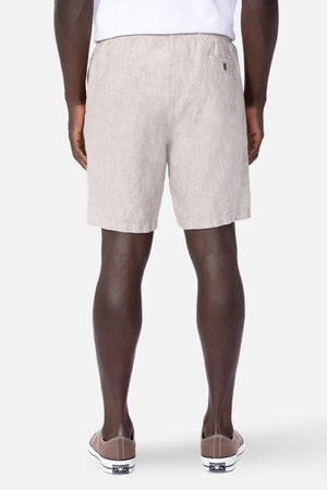 A man stands with his back to the camera in a white shirt, taupe slip-on sneakers, and Industrie The Marina Linen Short—light beige shorts celebrated for their relaxed, breathable fit. The background is plain and light gray.