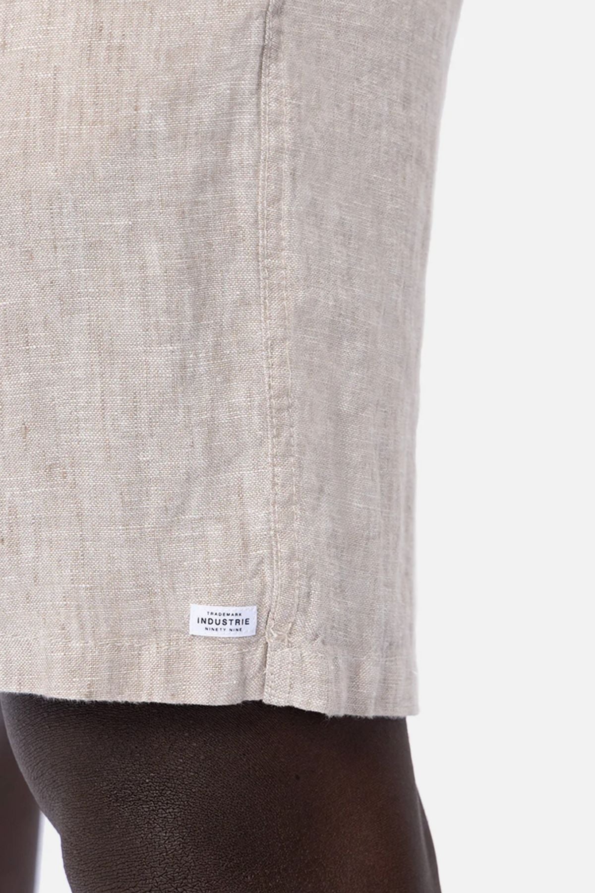 Close-up of a person wearing Industrie The Marina Linen Short in light beige with a small white "Industrie" label near the hem, set against a plain white background.