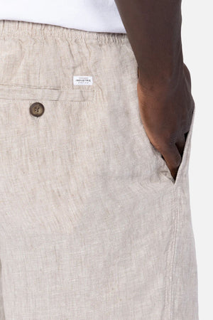A close-up of a person wearing Industrie The Marina Linen Short in light beige, with breathable fabric, relaxed fit, a buttoned back pocket, and a small white brand label above the pocket. Their right hand rests in the pocket.