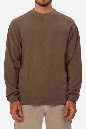 A man stands against a plain white background wearing Industrie The Marlon Long Sleeve Tee—a loose-fitting, 100% cotton brown crewneck—paired with light-colored pants.