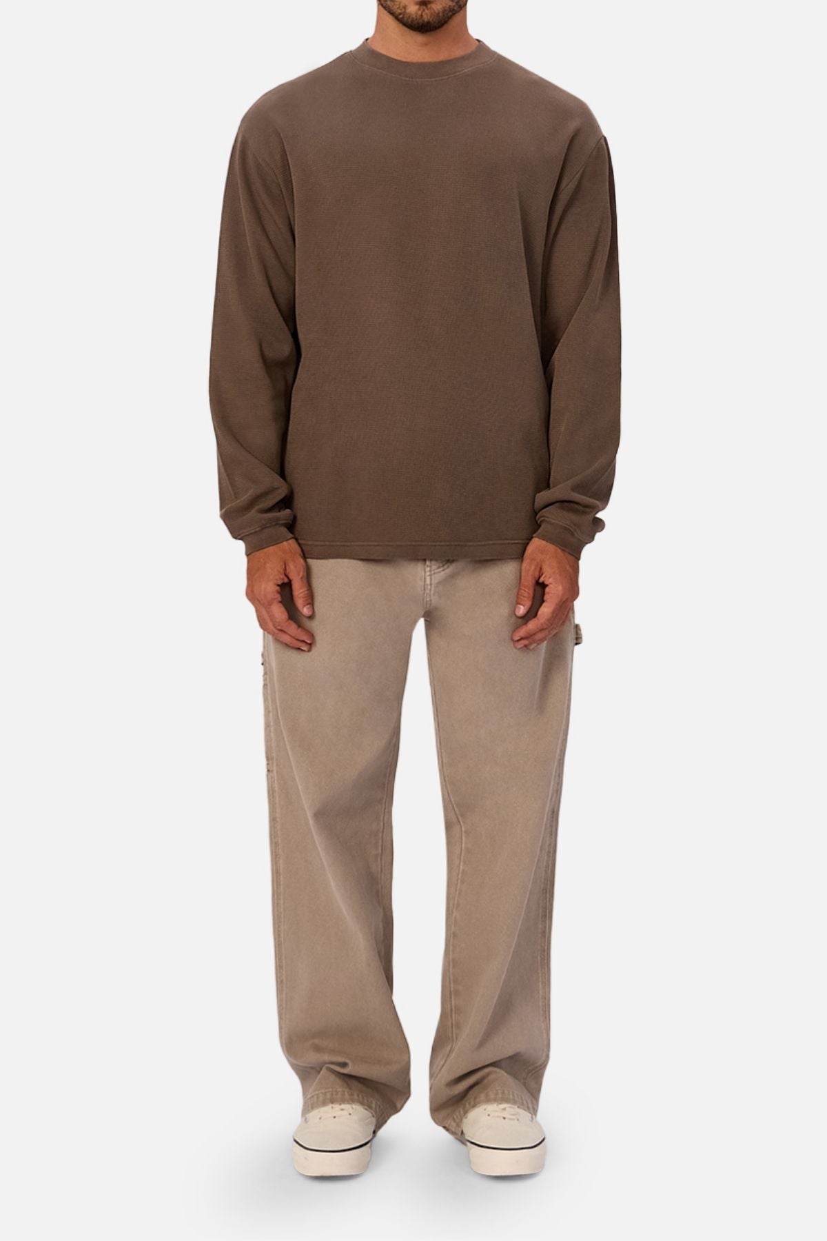 A person stands facing forward, dressed in the Industrie The Marlon Long Sleeve Tee in brown—made from 100% cotton—with light beige wide-leg jeans and white sneakers, set against a plain white background.