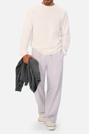 A person in a white sweatshirt, light gray sweatpants, and white sneakers holds an Industrie The Marlon Long Sleeve Tee, crafted from 100% cotton textured fabric, in dark gray with their left hand against a plain light background.