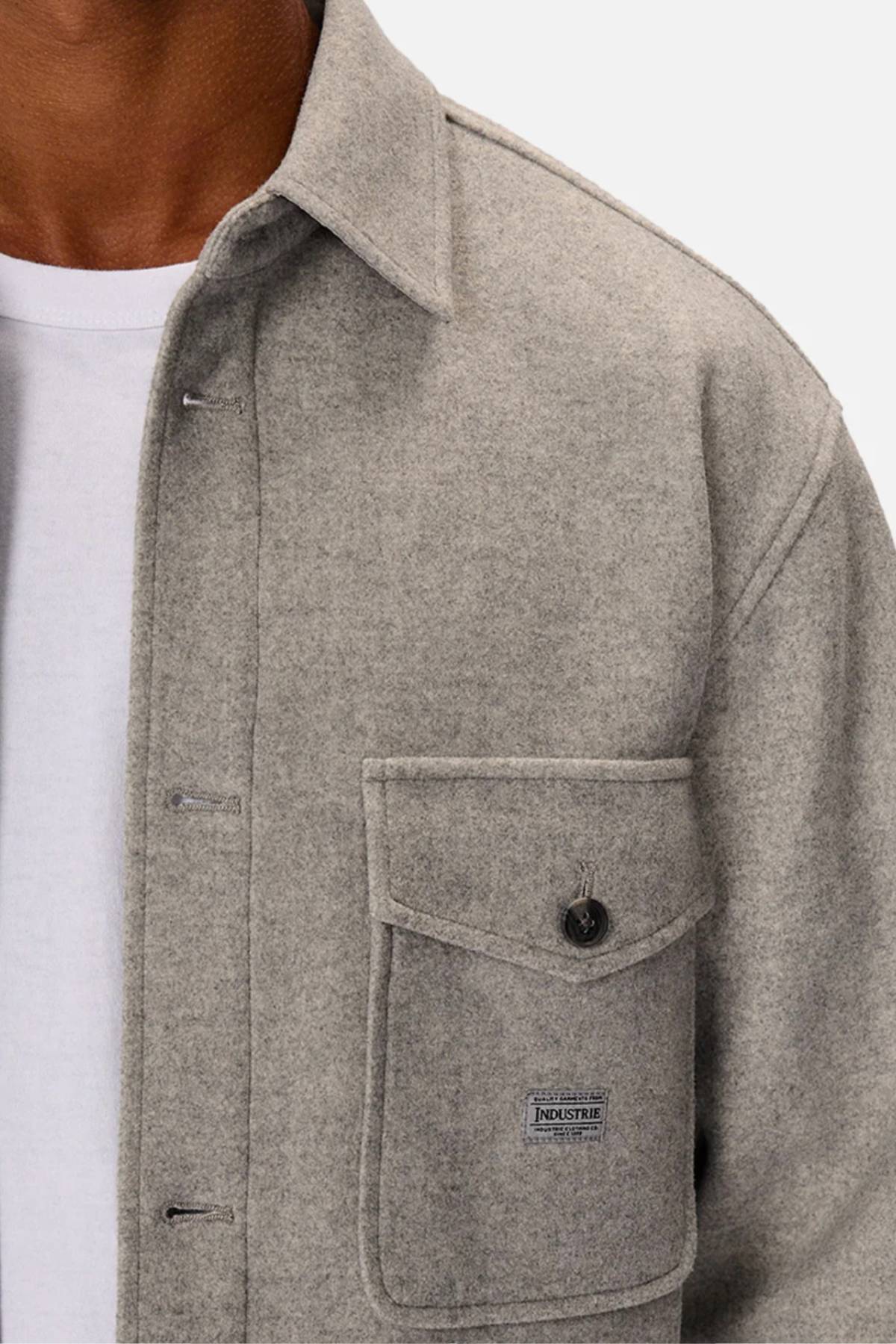 A person wears the Industrie The New Coleman Jacket—a light gray, relaxed fit wool shirt jacket with a classic collar—over a white t-shirt. It features a chest flap pocket with button and an "INDUSTRIE" logo tag.